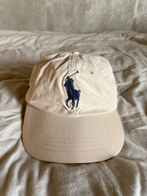 Beige keps från Polo Ralph Lauren - Snygg beige keps från Polo Ralph Lauren med klassisk mörkblå polospelare broderad framtill och siffran 3 på sidan. Justerbar baktill och tillverkad i bomull för en bekväm passform. Perfekt accessoar för en avslappnad och stilren look.