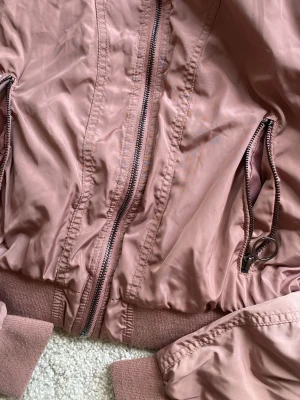 Rosa bomberjacka med dragkedja - Snygg rosa bomberjacka i glansigt material med ribbade muddar vid ärmslut, krage och nederkant. Jackan har två fickor med dragkedja och en klassisk bomberjacke-passform. Perfekt för dig som vill ha en stilren och trendig look. Passar M, L