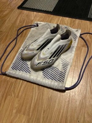  Adidas F50 fotbollsskor  - Säljer ett par vita Adidas F50 fotbollsskor med svarta och guldiga detaljer. Skorna har en strömlinjeformad design utan snörning och tydliga F50- och Adidas-loggor på ovansidan. Perfekta för dig som vill ha lätta och snabba skor på planen.