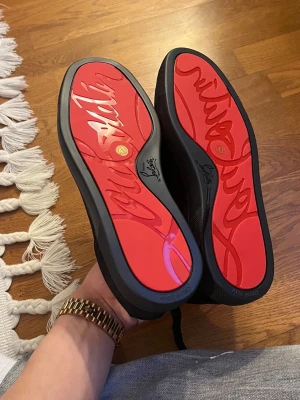 Christian Louboutin - Säljer ett par svarta sneakers från Christian Louboutin med ikonisk röd sula och signaturdetaljer undertill. Skorna har en clean och stilren design i svart mocka och textil, med svarta skosnören och diskreta hål för ventilation. Perfekta för dig som gillar exklusiva detaljer. Använda 1-2 gånger
