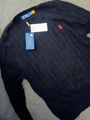 Svar stickad tröja Polo Ralph Lauren - Mörkblå stickad tröja från Polo Ralph Lauren med klassisk kabelstickning och rosa broderad logga på bröstet. Tröjan har rund halsringning och långa ärmar. Perfekt för en stilren och avslappnad look.