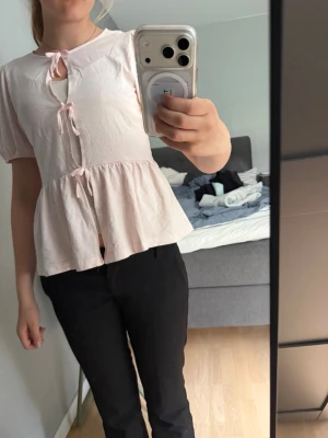 Ljusrosa blus med rosetter - Supersöt ljusrosa blus med korta puffärmar och tre dekorativa rosetter framtill. Blusen har en lätt utsvängd peplum-modell och är gjord i ett tunt, mjukt tyg som känns luftigt. Perfekt för dig som gillar en romantisk och feminin stil.