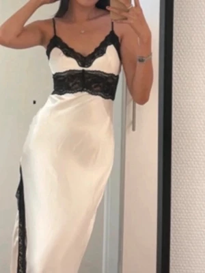 Vit långklänning med svart spets - Säljer en elegant vit långklänning med tunna axelband och v-ringad urringning. Klänningen har svarta spetsdetaljer vid bysten och i sidan, samt en slits som ger en snygg touch. Materialet ser ut att vara satin eller liknande för en lyxig känsla.