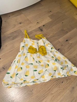 Vit och gul klänning med rosett - Söt kortklänning i vitt med gult citrusrandigt mönster och stora gula rosetter framtill. Klänningen har smala axelband och en luftig passform, perfekt för varma dagar. Materialet känns som bomull och ger en lätt och bekväm känsla.