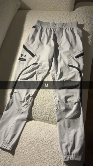 Grå mjukisbyxor från Under Armour - Säljer ett par grå mjukisbyxor från Under Armour med svarta detaljer och dragkedjor vid fickorna. Byxorna har resår i midjan och vid bensluten, samt coola sömmar och sportig look. Perfekta för chill eller träning.