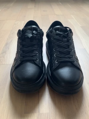 Svarta sneakers från Karl Lagerfeld - Stilrena helsvarta sneakers från Karl Lagerfeld med lågt skaft och klassisk snörning. Skorna har en diskret logga på sidan och är tillverkade i slätt läder med detaljer i textil. Perfekta för dig som gillar en clean och modern look. Storlek 42