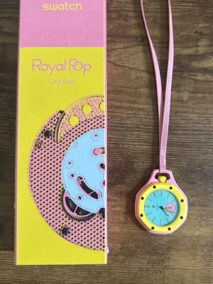 Swatch Royal Pop OTG Roz klocka - Unik Swatch Royal Pop OTG Roz klocka med färgglad design i gult, rosa och blått. Klockan har ett runt ur med grafiskt mönster och hängs i ett rosa band. Perfekt accessoar för dig som gillar lekfull stil och poppiga färger.
