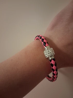 Flätat rosa och svart armband - Snyggt armband med flätad design i rosa och svart. Armbandet har ett silverfärgat pärl-lås dekorerat med små glittrande stenar. Perfekt accessoar för att ge din outfit en färgklick och lite extra bling. Magnetlås