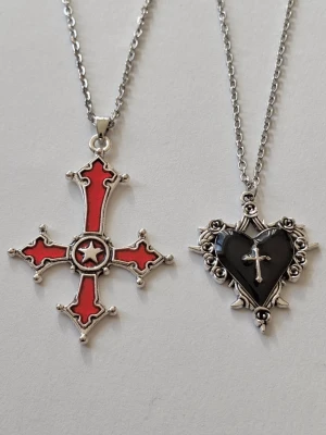 Två halsband med goth och hjärta - Säljer två halsband i silverfärgat rostfitt stål. Ena är kors med röd emalj och stjärndetalj i mitten, den andra har ett svart hjärta med svart emalj och små rosor, taggar runt och ett litet kors ovanpå. Båda hängs på tunna kedjor och har en tydlig gotisk vibe.