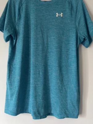 Blått träningsset från Under Armour - Snyggt träningsset med en blå t-shirt och matchande blå shorts från Under Armour. T-shirten har rund hals och logga på bröstet, shortsen har resår i midjan och logga längs benet. Perfekt för gym eller löpning.