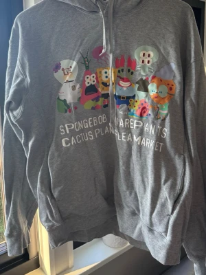 Uniqlo x Cactus Plant Flea Market x Spongebob - Säljer en grå hoodie från Uniqlo, CPFM med färgglatt Spongebob Squarepants-tryck i samarbete med Cactus Plant Flea Market. Hoodien har huva med dragsko och känguruficka framtill. Storlek Medium.  Perfekt för dig som gillar streetwear och unika samarbeten.