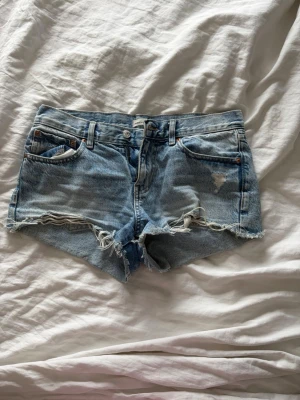 Ljusblå jeansshorts med fransig kant - Säljer ett par ljusblå jeansshorts med fransig, rå kant och slitningar framtill. Klassisk femficksmodell med bälteshällor och knappgylf. Perfekta för varma dagar och en avslappnad stil.