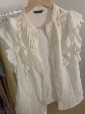 Vit zara blus  - Söt vit blus i luftig bomull med volangdetaljer längs axlarna och fina veckade partier framtill. Blusen är ärmlös och har rund halsringning samt knäppning framtill. 