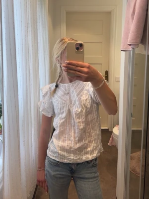 Vit blus med volang från Forever New - Söt vit blus från Forever New med volangdetaljer och spets vid axlarna. Blusen har korta ärmar och ett tunt, luftigt tyg med diskreta ränder. Perfekt till jeans eller kjol för en fräsch och somrig look.