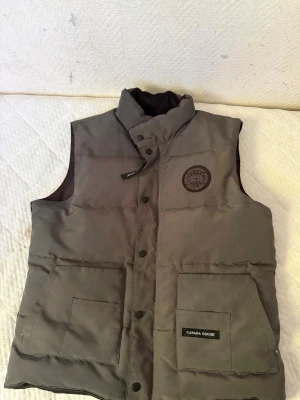Grå dunväst från Canada Goose - Snygg grå dunväst från Canada Goose med hög krage och två stora fickor framtill. Västen har tryckknappar och ett broderat märke på bröstet. Perfekt för lager-på-lager och håller dig varm under kyliga dagar.