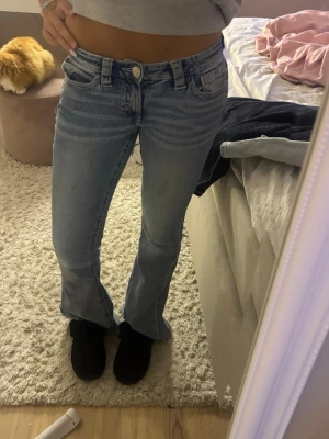 Ljusblå bootcut jeans med detaljer - Säljer ett par ljusblå bootcut jeans med snygga slitningar och dekorativa sömmar på bakfickorna. Jeansen har låg midja och klassisk femficksdesign. Perfekta för dig som gillar en avslappnad men trendig stil.