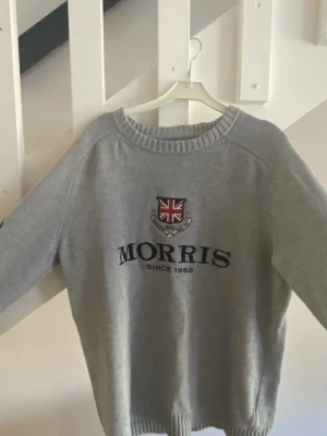 Grå sweatshirt från Morris - Grå sweatshirt från Morris med broderad logga och brittisk flagga på bröstet. Tröjan har rund halsringning, långa ärmar och ribbade muddar. Perfekt för en avslappnad och stilren look.