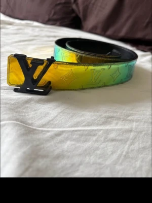 Revirseable Louis Vuitton bälte - Säljer ett Louis Vuitton bälte med stor LV-logga i svart metall. Bältet har en cool gradient i gult, grönt och blått med klassiskt LV-mönster. Perfekt statement piece för att lyfta vilken outfit som helst.