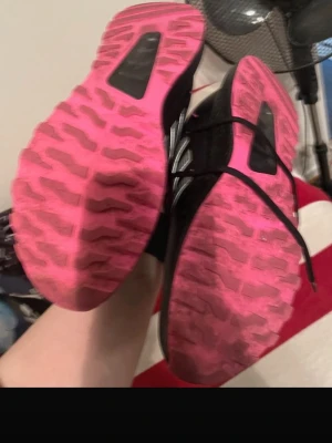Svarta Adidas sneakers med rosa sula - Snygga svarta Adidas sneakers med klassiska vita ränder på sidan och rosa detaljer på insidan samt yttersulan. Skorna har snörning och en sportig look med greppvänlig sula i rosa. Perfekta för dig som gillar streetstyle och sportig vibe.