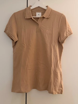 Pikétröja från BMW - Snygg beige pikétröja från BMW med klassisk krage och korta ärmar. Tröjan har diskret broderad BMW-logga på bröstet och är tillverkad i ett bekvämt bomullsmaterial. Perfekt för en stilren och avslappnad look.