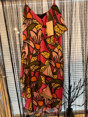 Färgglad blommig omlottklänning - Säljer en superfin kortklänning med stort blommönster i rosa, gul, orange och grönt. Klänningen har omlottdesign, volanger och tunna axelband. Perfekt för dig som vill sticka ut med färg och mönster!