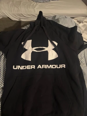 Svart hoodie från Under Armour - Svart hoodie från Under Armour med stor vit logga och text på bröstet. Klassisk modell med huva och dragsnören samt en stor magficka. Perfekt för dig som gillar sportig och avslappnad stil. Det är mest till träning så den är lite lite större u storleken 