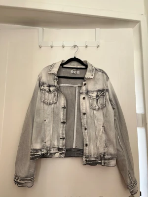 Jeansjacka - Snygg grå jeansjacka med tvättad effekt och svarta kontrastsömmar. Klassisk modell med två bröstfickor med lock och knappar framtill. Jackan har en avslappnad passform och justerbara knappar i midjan. 