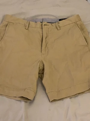 Beige shorts från Polo Ralph Lauren - Klassiska beige shorts från Polo Ralph Lauren med normal passform. Tillverkade i mjuk bomull och har bälteshällor, sidofickor och knappgylf. Perfekta för varma dagar och enkel att matcha med olika stilar.