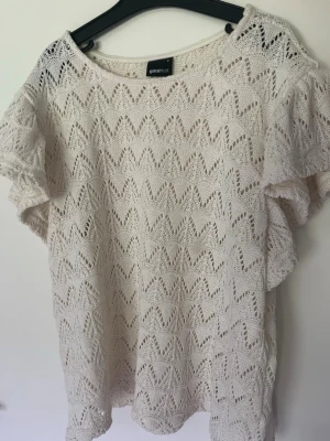 Vit topp från Gina tricot - Supersöt vit topp från Gina Tricot i spetsstickat material med korta ärmar och rund hals. Mönstret är genombrutet i zigzag och ger en luftig känsla. Perfekt till jeans eller kjol för en fräsch look.