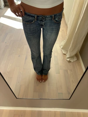 Blå bootcut jeans med nitar - Säljer ett par blå bootcut jeans med slitningar och snygga nitar på fickorna. Jeansen har låg midja och klassisk femficksmodell. Perfekta för dig som gillar en avslappnad men trendig stil.