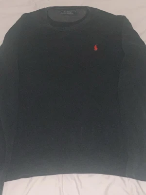 Mörkblå stickad tröja från Polo Ralph Lauren - Klassisk mörkblå stickad tröja från Polo Ralph Lauren i mjuk pima bomull. Tröjan har rund halsringning och den ikoniska röda logotypen broderad på bröstet. Perfekt till jeans eller chinos för en clean och stilren look.