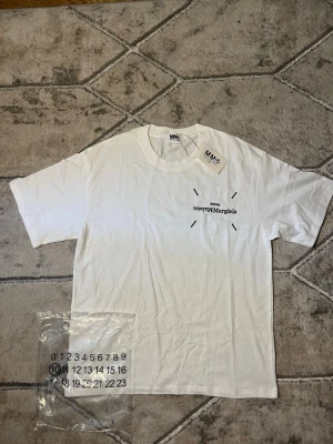 Vit t-shirt från Maison Margiela - Vit t-shirt från Maison Margiela med svart tryck på bröstet och klassisk rund hals. T-shirten har korta ärmar och är gjord i mjuk bomull. En stilren och trendig tee som passar perfekt till jeans eller shorts.