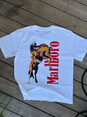 Vit Marlboro t-shirt med cowboytryck vintage - Storlek S 100%bomull bra kvalitets tshirt. Unisex Vit t-shirt med stort tryck av en cowboy på häst och röd Marlboro-text på ryggen. Klassisk passform och korta ärmar, perfekt för dig som gillar westernstil och statement-plagg.