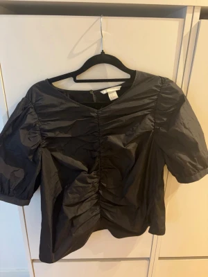 Svart puffärm blus med rynk - Snygg svart blus med puffiga korta ärmar och rynkdetalj framtill. Blusen har rund halsringning och är tillverkad i ett glansigt syntetmaterial som ger en cool look. Perfekt för dig som vill ha något trendigt och stilrent.