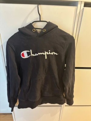 Svart Champion hoodie med logga - Snygg svart hoodie från Champion med stor broderad logga på bröstet och en mindre logga på ärmen. Klassisk känguruficka framtill och huva med snörning. Perfekt för en chill och sportig look. Mjuk bomullsblandning som är skön att ha på sig.