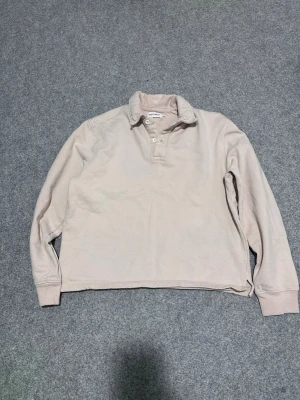 Beige långärmad pikétröja - Säljer en beige långärmad pikétröja med krage och knappar framtill. Tröjan har en avslappnad passform och ribbade muddar vid ärmsluten. Perfekt för en clean och enkel stil.