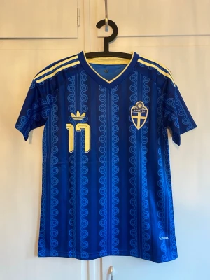 Blå Sverige fotbollströja Adidas - Säljer en blå fotbollströja från Adidas med Sveriges landslagsemblem och nummer 17 i gult. Tröjan har GYÖKERES tryckt på ryggen och klassiska Adidas-ränder på axlarna. Materialet är lätt och andas, perfekt för träning eller match.