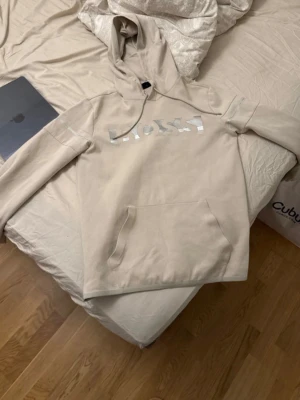 Beige boss hoodie med silver BOSS-logga - Säljer en beige hoodie med stor BOSS-logga i vitt och silver på bröstet. Hoodien har huva med dragsko och en stor magficka. Tillverkad i mjukt bomullsmaterial och har en avslappnad passform, perfekt för en chill och stilren look.