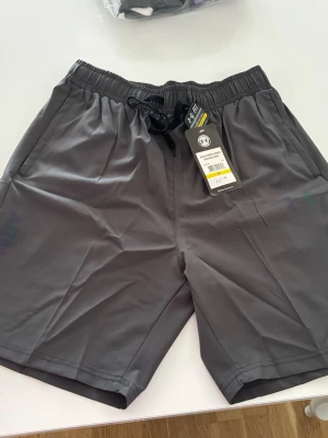 Grå träningsshorts  - Snygga grå träningsshorts från Under armor med elastisk midja och snörning. Shortsen har en logga på benet och är tillverkade i ett lätt och snabbtorkande syntetmaterial. Byxorna kommer i stl M men passar S ännu bättre.