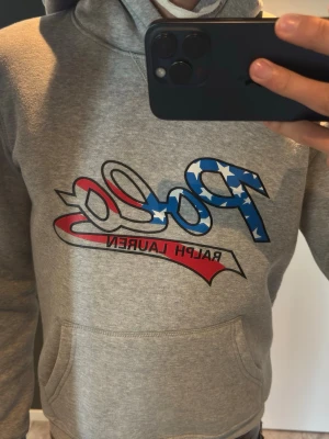 Grå hoodie från Polo Ralph Lauren - Grå hoodie med stor magficka och dragsko i huvan. Framsidan har Polo Ralph Lauren-logga med amerikanska flaggans färger och stjärnor. Mjuk bomullskänsla och klassisk passform, perfekt för en avslappnad stil.