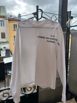 Vit långärmad Supreme x Comme des Garçons tee - Säljer en clean vit långärmad t-shirt från Supreme i collab med Comme des Garçons. Tryck i svart text på bröstet och klassisk rund hals. Perfekt för dig som gillar streetwear och vill sticka ut med en limited edition tee.