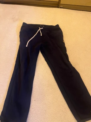 Mörkblå mjukisbyxor från Polo Ralph Lauren - Säljer ett par mörkblå mjukisbyxor från Polo Ralph Lauren med gul broderad logga på benet och vit dragsko i midjan. Byxorna har avslappnad passform och är perfekta för chill dagar. Tillverkade i mjukt material för extra komfort.