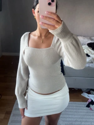 Beige stickad tröja med fyrkantig ringning - Snygg beige stickad tröja med lång ärm och trendig fyrkantig ringning. Tröjan har ribbade muddar och sitter tajt över kroppen, perfekt för en stilren look. Passar till både jeans och kjol för en modern outfit.