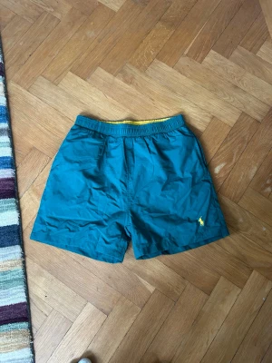  Polo Ralph Lauren badbyxor - Snygga grönturkosa badbyxor från Polo Ralph Lauren med gul logga på benet och gul insida i midjan. Klassisk modell med resår i midjan och meshfoder för extra komfort. Perfekta för stranden eller poolen.