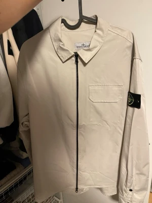 Beige overshirt från Stone Island - Snygg beige overshirt från Stone Island med dragkedja framtill, klassisk krage och bröstficka. Ikoniska Stone Island-märket på ärmen. Tillverkad i ett lätt material som passar perfekt för lager-på-lager-stil.
