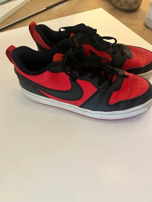 Nike svart-röda låga sneakers - Säljer ett par klassiska Nike sneakers i svart och rött med lågt skaft. Skorna har svart swoosh, svarta skosnören och vit sula. Ovandelen är i läder med perforerade detaljer på tån för extra ventilation. Perfekta för en streetwear-look.