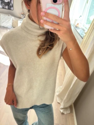 Beige stickad väst med polokrage - Säljer en beige stickad väst med hög polokrage och breda ärmhål. Västen har en avslappnad passform och är perfekt att styla över en skjorta eller t-shirt. Materialet är mjukt och ger en mysig känsla, perfekt för lager på lager under kyligare dagar.