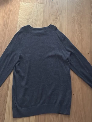 Mörkgrå stickad tröja - En enkel och stilren mörkgrå stickad tröja med rund halsringning och långa ärmar. Tröjan har en rak passform och ribbade muddar vid ärmslut och nederkant. Perfekt basplagg till garderoben.