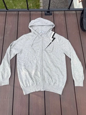Grå hoodie med blixtdetalj - Snygg grå hoodie med dragkedja från Baldy. Hoodien har en cool blixtdetalj i svart och vitt på ena axeln, dragsko i huvan och ribbade muddar. Tillverkad i mjukt material som känns skönt mot huden. Perfekt för en avslappnad och trendig look.