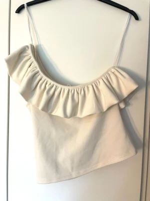 Vit topp med volang - Supersöt vit topp med volang längs urringningen. Det är en one-shoulder topp. Perfekt för varma dagar och enkel att styla med jeans eller kjol. Defekt: hål i ena sidan men syns inte väl när den e på💕